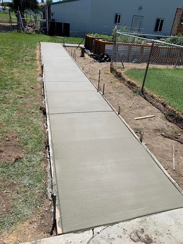 Newly poured concrete pathway with wooden forms along a residential yard, metal fence visible nearby